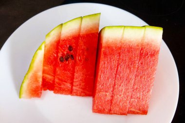 Lezzetli, taze karpuz dilimleri beyaz bir tabakta düzgünce dizilmiş. Canlı kırmızı ve yeşil renkler sağlıklı beslenme temaları, yemek fotoğrafçılığı, yaz içeceği, aşçılık dergileri ve görsel olarak çekici içerik arayan tarif blogları için ideal hale getiriyor..