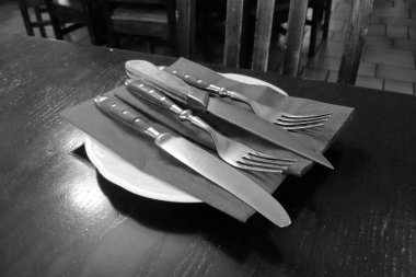 Beyaz bir tabağın üzerine katlanmış bir peçeteye düzgünce yerleştirilmiş çatal ve bıçaklarla sade bir restoran masasının siyah beyaz fotoğrafı. Arkadaki ahşap sandalyeler ve fayanslı zemin rahat bir yemek atmosferi olduğunu gösteriyor..
