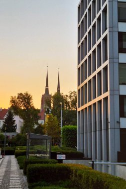 Wroclaw, Polonya, 23 Ağustos 2024: Günbatımındaki tarihi kilise kuleleriyle modern mimari çelişiyor. Sahnede yeşil alanlar boyunca çağdaş binalar, doğa ile şehir yaşamını harmanlarken, akşam gökyüzü parlıyor.