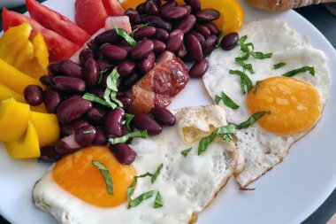 Canlı bir kahvaltı tabağı. İçinde güneşli yumurta, fasulye, taze domates, sarı dolmalık biber ve yapraklı yeşillikler var. Besleyici bir başlangıç için mükemmel, renkli ve dengeli bir yemek seçeneği sunuyor.