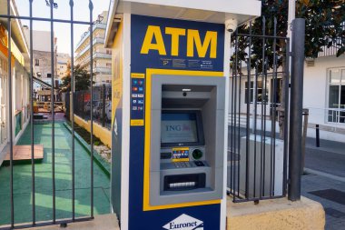 Lloret de Mar, İspanya, 3 Ekim 2024: Güvenliği için metal çitlerle çevrili, hareketli bir şehir sokağında bulunan bir açık hava bankası. Sahne, önemli bir bankamatik görüntüsü ve şehir mimarisi ile kentsel mali işlemlerde kolaylığı vurguluyor