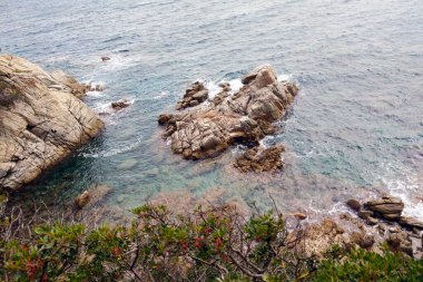 Engebeli kayaların kristal berrak sulara girdiği sakin sahil manzarası. Canlı yeşillik manzarayı çerçevelerken, nazik dalgalar kıyı şeridine doğru kıvrılır. Doğa aşıkları ve seyahat meraklıları için mükemmel..