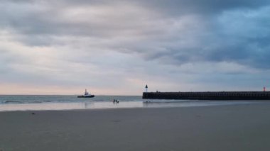 Sahilde sakin bir akşam. Deniz fenerine yaklaşan bir römorkör var. Geniş kumlu sahil dramatik bir gökyüzü altında sakin bir okyanusla buluşuyor, sakin kıyı atmosferi..