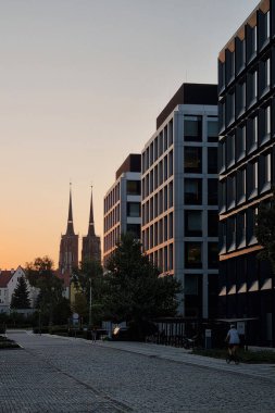 Wroclaw, Polonya, 23 Ağustos 2024: Günbatımında sakin bir şehir manzarası modern ofis binaları ve uzaktaki bir katedralin çarpıcı kuleleri. Mimariyle doğanın ahenkli karışımı günden güne barışçıl geçişi vurguluyor. 