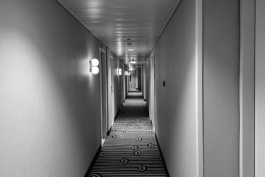 Modern iç tasarımın yer aldığı bir otel koridorunun siyah beyaz fotoğrafı. Koridorda geometrik halı desenleri ve duvar apliklerinden gelen yumuşak ortam aydınlatmaları sergileniyor. Minimalist ve zarif bir atmosfer yaratıyorlar..
