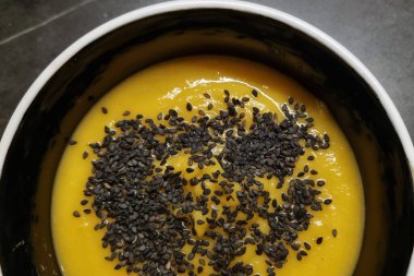 Siyah susam tohumlarıyla kaplı sıcak, kremalı balkabağı çorbası mükemmel bir lezzet ve doku karışımı sağlıyor. Sıcak ve sağlıklı yemekler için ideal. Mutfak fotoğrafçılığında konfor ve beslenme özünü yakalar..