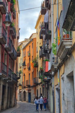 Girona, İspanya, 3 Ekim 2024: Avrupai bir kasabada, balkon ve bitkilerle süslenmiş canlı binalarla dolu pitoresk dar bir sokak. Ön cephelerden bayraklar sarkıyor ve olay yerinden geçen kaldırım taşı yolu tuhaf atmosferi güçlendiriyor.