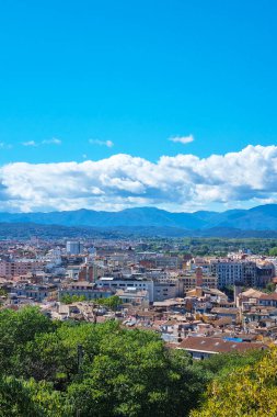 Girona, İspanya, 3 Ekim 2024: Canlı, mavi bir gökyüzü altında uzak dağ arkaplanlarına karşı kentsel mimarinin bir karışımını içeren şehir manzarası. Seyahat ve turizm promosyonları, şehir hayatı ve doğal güzellik için ideal.