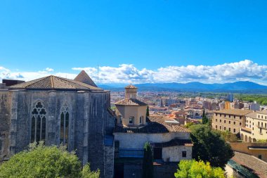 Girona, İspanya, 3 Ekim 2024: Gotik tarzı elementlere sahip tarihi binalar açık mavi gökyüzünün altındaki canlı bir şehir manzarasını görmezden geliyor. Yeşillik antik mimariyi tamamlıyor ufuktaki uzak dağlar dinginliği arttırıyor, pictu