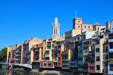 Girona, İspanya, 3 Ekim 2024: Onyar Nehri parlak mavi bir gökyüzünün altında Girona, İspanya 'da sıralanır. Arkadan çarpıcı bir gotik katedral kulesi yükseliyor ve şehrin canlı mimari cazibesini arttırıyor..