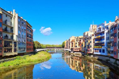 Girona, İspanya, 3 Ekim 2024: Renkli cepheli binalar nehrin her iki yakasında da yankılanıyor, yansımaları aşağıdaki durgun suda yansıyor. Küçük bir köprü iki tarafı tek bir bulutla mavi bir gökyüzünün altında birbirine bağlar..