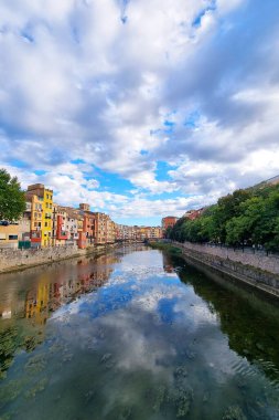 Girona, İspanya, 5 Ekim 2024: Canlı mimari dramatik bir gökyüzü altında sakin bir nehir oluşturur. Yansımalar, seyahat temaları, mimari çalışmalar veya Avrupa manzarası özellikleri için ideal sakin bir sahne oluşturur.