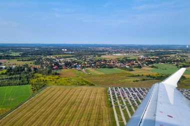 Nefes kesici bir hava fotoğrafı, açık mavi bir gökyüzü altında geniş alanlar, kompakt bir kasaba ve uzak dağlar sergilenen, bir uçak penceresinden görülen bir manzara yakalar. Görüntü, kırsal ve kentsel alanlar arasındaki karşıtlığı vurgular.