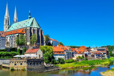 Goerlitz, Almanya, 6 Temmuz 2024: Huzurlu bir nehir boyunca büyük bir gotik katedralin ve büyüleyici kırmızı çatılı binaların yer aldığı tarihi bir kasabanın manzarası. Canlı yeşillik ve berrak mavi gökyüzü seyahat için ideal olan resim ortamını güçlendirir.