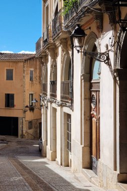 Girona, İspanya, 3 Ekim 2024: Güneş ışığı, tarihi Akdeniz binalarıyla çevrili dar bir kaldırım taşı caddesini aydınlatıyor. Klasik mimaride süslü balkonlar, kemerli kapılar ve dekoratif fenerler var. Resimli bir şehir yaratıyor.