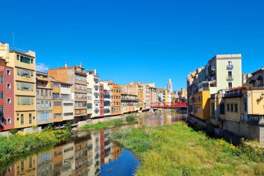 Girona, İspanya, 3 Ekim 2024: Açık mavi bir gökyüzünün altında Girona, İspanya 'da nehir kıyıları boyunca uzanan renkli binalar. İkonik kırmızı köprü nehir yolu boyunca canlı mimariyi ve yemyeşil yeşilliği yansıtan su yolunu kapsar..