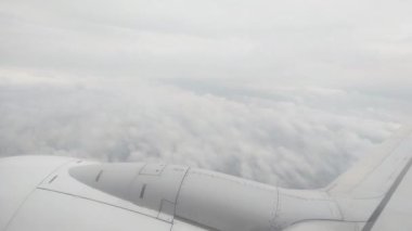 Ufuk boyunca uzanan kabarık bulutları gösteren bir uçak penceresinden bak. Uçak kanadı görülebilir, bulutların üzerinde uçarken huzurlu ve huzurlu bir perspektif sunuyor.
