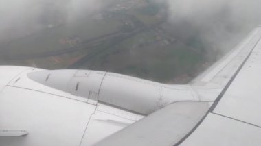 Uçuşun ortasındaki bir uçağın manzarası, uçağın kanadının altında kısmen bulutlu gökyüzünden görünen manzaralı arazi parçalarını gösteriyor. Perspektif, havacılık seyahatini ve altındaki geniş manzarayı vurgular..