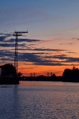 Sakin bir nehrin üzerinde gün batımının olduğu sakin bir akşam sahnesi. Renkli gökyüzüne karşı siluetlenmiş bir köprü ve yüksek bir iletim kulesi, doğal ve insan yapımı elementlerin uyumlu bir karışımını oluşturuyor..