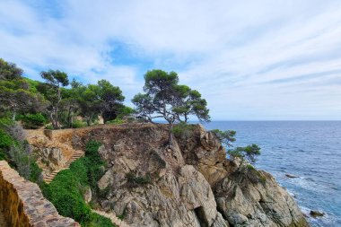Çam ağaçlarıyla süslenmiş kayalık bir uçurum yolu bulunan kıyı manzarası. Huzurlu Akdeniz, parçalı bulutlu bir gökyüzünün altında ufka doğru uzanıyor. Huzurlu ve resimsel bir manzara yaratıyor..