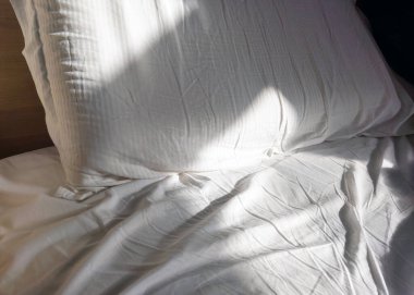 Sabah güneşi, rahat beyaz çarşaflar ve yastıklarla ıssız bir yatağa akarak sakin ve sakin bir atmosfer yaratır. Sahne, rahatlama, konfor ve ev sakinliğini çağrıştırıyor. Yaşam tarzı ve iç tasarım temaları için mükemmel..