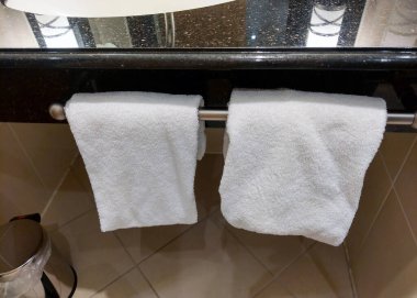 Modern bir banyo sahnesinde parlak bir tezgahın altında parlak metalik bir askıda asılı iki peluş beyaz havlu görülüyor. Şık düzenleme, çağdaş bir ortamda temizlik ve lüks hissi sunuyor..