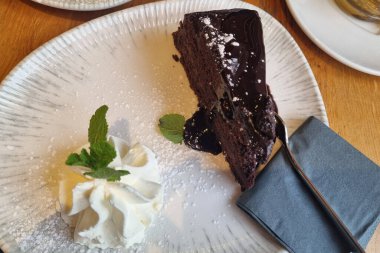 Çikolata soslu, nane yaprağıyla süslenmiş bir dilim zengin çikolatalı pasta ve yanında da bir tutam krem şanti. Beyaz seramik bir tabağa peçete ve kaşıkla nazikçe yerleştirilmiş..