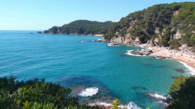 Turkuaz suların, tenha bir plajın ve açık mavi gökyüzünün altındaki yeşil yamaçların yer aldığı çarpıcı sahil manzarası. Seyahat broşürleri ve doğa temalı tasarımlar için mükemmel, dinginliği çağrıştırıyor ve