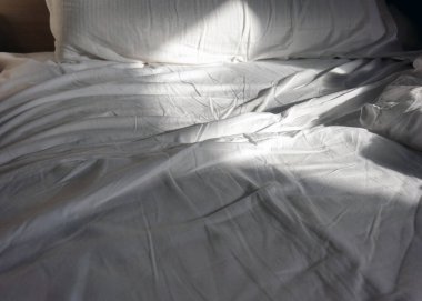 Güneş ışığı beyaz yatak çarşaflarını nazikçe aydınlatır ve sakin bir atmosfer yaratır. Buruşuk kumaşın dokusu sıcaklık ve rahatlık ekler. Rahatlık ve sakinlik hissi uyandırır..