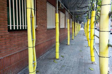 Sarı güvenlik bariyerleri ve kırmızı tuğlalı bina boyunca iskele olan bir kaldırım manzarası. Koruyucu kaplamaların düzenli olarak hizalanması kentsel gelişimi ve yaya güvenliğini vurguluyor. Düşen yapraklar doğaya dokunur..