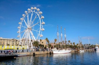 Barselona, İspanya, 25 Aralık 2024: Büyük bir dönme dolap ve uzun yelkenli gemilerin yer aldığı liman sahnesi hareketli bir gezinti güvertesine kenetlendi. açık mavi gökyüzü ve palmiye ağaçları bölgenin canlı atmosferini arttırır..