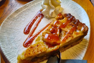 Seramik tabağa yerleştirilmiş cazip bir karamelli elmalı turta dilimi. Tatlı üzerine karamel sosu serpilmiş ve yanında bir tutam krema ile tatlı aşıkları için mükemmel bir ziyafet sunuluyor..