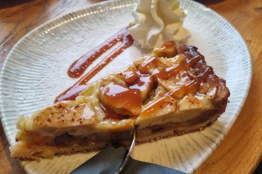 Üzerinde bol karamelli çiselemeli lezzetli bir dilim elmalı turta ve yanında da bir parça krem şanti. Kırsal seramik tabakta servis edilen turta vitrinleri altın-kahverengi kabuk ve yumuşak elma dolgusu sıcak, davetkar bir tatlı ortamı yaratıyor..