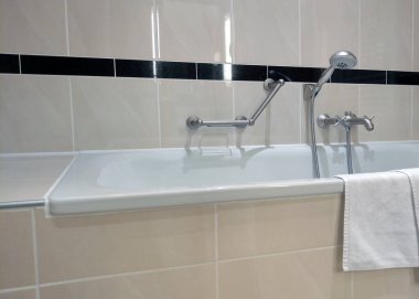 Modern krom armatürleri olan parlak beyaz bir küvetin olduğu rahat bir banyo. Duvarlar ve zemin parlak bej fayanslarla süslenmiş, şık bir siyah fayans aksanıyla tamamlanmış. Beyaz bir havlu düzgünce asılır ve biraz rahatlık katar.