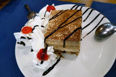 Çikolata sosuyla süslenmiş, üzerinde tiramisu tozu bulunan nefis bir dilim beyaz bir tabakta duruyor. Yanında çırpılmış krema ve canlı kirazlar ile birlikte, tatlı mavi bir masa örtüsüne karşı ayarlanmış, görsel bir... 