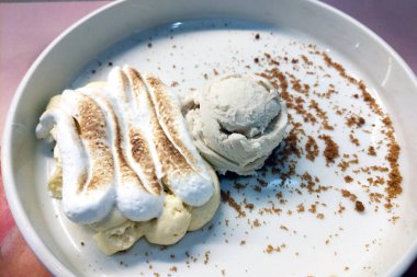 Bir kepçe dondurma ve bol miktarda kızarmış tepeli krema içeren nefis bir tatlı. ustalıkla parçalanmış bir parçayla süslenmiş bu yemek zarif bir şekilde yuvarlak beyaz bir tabakta sunuluyor..