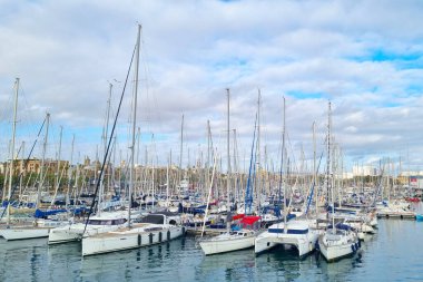 Barselona, İspanya, 25 Aralık 2024: Parçalı bulutlu bir gökyüzü ve uzak kentsel gökyüzü tarafından çerçevelenmiş hareketli bir marinayı yelkenliler ve yatlar dolduruyor. Huzurlu su damarları yansıtır, sakin ama dinamik bir denizcilik ortamı yaratır yolculuk ve yolculuk için ideal