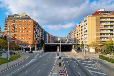 Barselona, İspanya, 1 Ocak 2025: Parlak mavi gökyüzü altında simetrik yerleşim alanlarının bulunduğu boş bir şehir caddesi. Yol, uzaktaki bir tünele çıkıyor. Ağaçlarla kaplı kaldırımlar ve trafik işaretleri şehrin sükunetini arttırıyor.