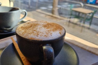 Üzerinde tarçın tozu olan dumanı tüten bir bardak kapuçino ve yanında tarçın çubuğu bir kafe masasında oturuyor. Bulanık arkaplan, rahat bir kafeterya ortamını işaret eden bir sokak görünümünü ortaya çıkarır..