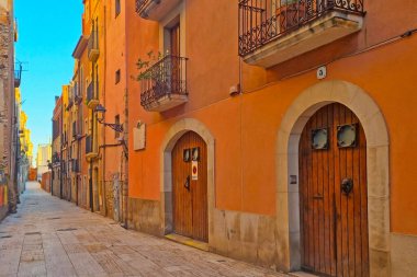 Tarragona, İspanya, 29 Aralık 2024: Dar bir Akdeniz caddesinin canlı ve renkli cephelerle kaplı manzarası. Geleneksel ahşap kapılar, süslü balkonlar, ve güneş ışığı alan, kaldırım taşı yolu sıcak ve davetkar bir atmosfer yaratır..