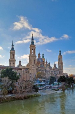Zaragoza, İspanya, 27 Aralık 2024: Bazilika del pilar 'ın çarpıcı manzarası, birden fazla kule ve kule ile barok mimarisini sergiliyor. Huzurlu Ebro nehri ön planda akar, güneş ışığını yansıtır, Zaragoz 'un ihtişamını artırır.