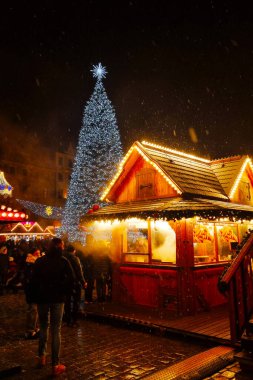 Wroclaw, Polonya, 5 Ocak 2025: Bir Noel pazarındaki şenlikli gece sahnesi, yüksek aydınlatılmış bir ağaç, sıcak ışıklandırmalı rahat ahşap tezgahlar ve yumuşak kar taneleri altında tatil atmosferinin keyfini çıkaran hareketli bir kalabalık.