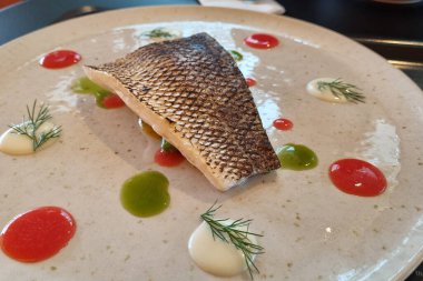 Seramik bir tabakta mükemmel pişmiş balık filetosu sunumu, canlı kırmızı, yeşil ve beyaz soslarla süslenmiş, taze dereotla süslenmiş, görsel olarak çekici bir mutfak başyapıtı yaratıyor..