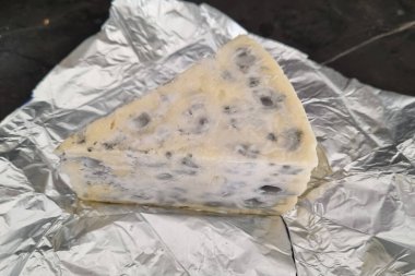 Buruşuk alüminyum folyoda, zengin bir dokusu ve kendine özgü kalıpları olan bir dilim mavi peynir yatıyor. Görüntü gurme peynirinin eşsiz özelliklerini vurgular, mutfak blogları, menüler veya yemek fotoğrafçılığı için idealdir.