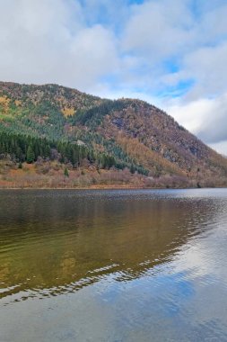 Sersemletici manzara, bulutlu bir gökyüzünün altında gür orman tepeleriyle çevrili berrak yansımalarıyla sakin bir gölü gösteriyor. Sakin su yüzeyi ve yeşillik huzurlu ve doğal bir ortam yaratır..