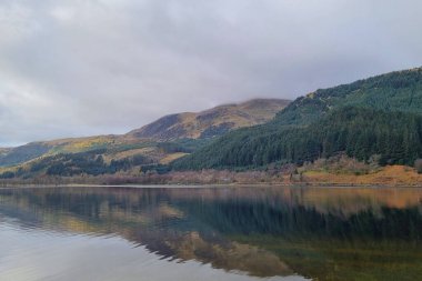 Huzurlu bir dağ manzarası, zümrüt çamlarla kaplı yamaçları ve engebeli tepeleri yansıtan durgun bir göle sahiptir. Bulutlu gökyüzü huzur dolu manzaraya derinlik katarak el değmemiş vahşi doğanın özünü ve huzurlu izolasyonu yakalıyor..