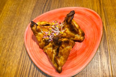 Üçgen şekilli hamantaschen hamur işleri, üzerinde narin filizler bulunan canlı kırmızı bir tabakta servis edilir. Altın kahverengisi, ahşap masa ile tezat oluşturarak aşçılık sunumları için iştah açıcı bir görsel yaratıyor..