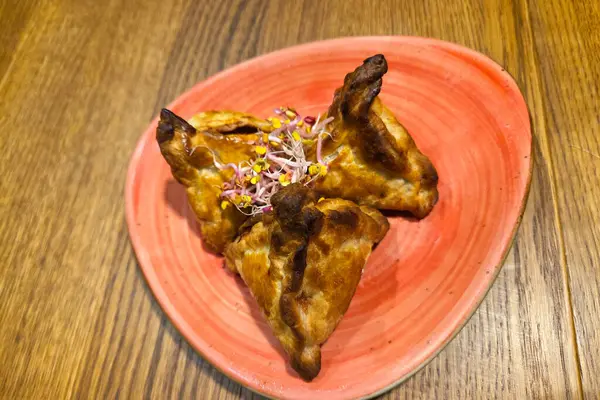 Üçgen şekilli hamantaschen hamur işleri, üzerinde narin filizler bulunan canlı kırmızı bir tabakta servis edilir. Altın kahverengisi, ahşap masa ile tezat oluşturarak aşçılık sunumları için iştah açıcı bir görsel yaratıyor..