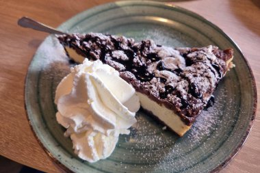 Çikolata soslu nefis bir dilim kremalı cheesecake, kırsal seramik tabakta bol miktarda krem şanti ile servis edilir, hoşgörü ve gurme tatlı estetiğini çağrıştırır..