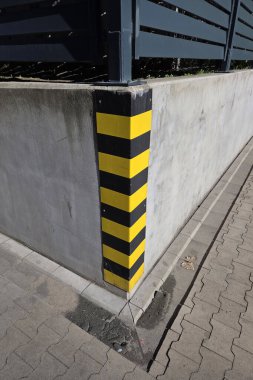 Kentsel güvenlik önlemlerinde beton bariyer üzerindeki siyah ve sarı uyarı çizgileri. Bariyer, metal bir çitle çevrili bir köşeyi koruyor. Şehir ortamında ihtiyatı simgeliyor..